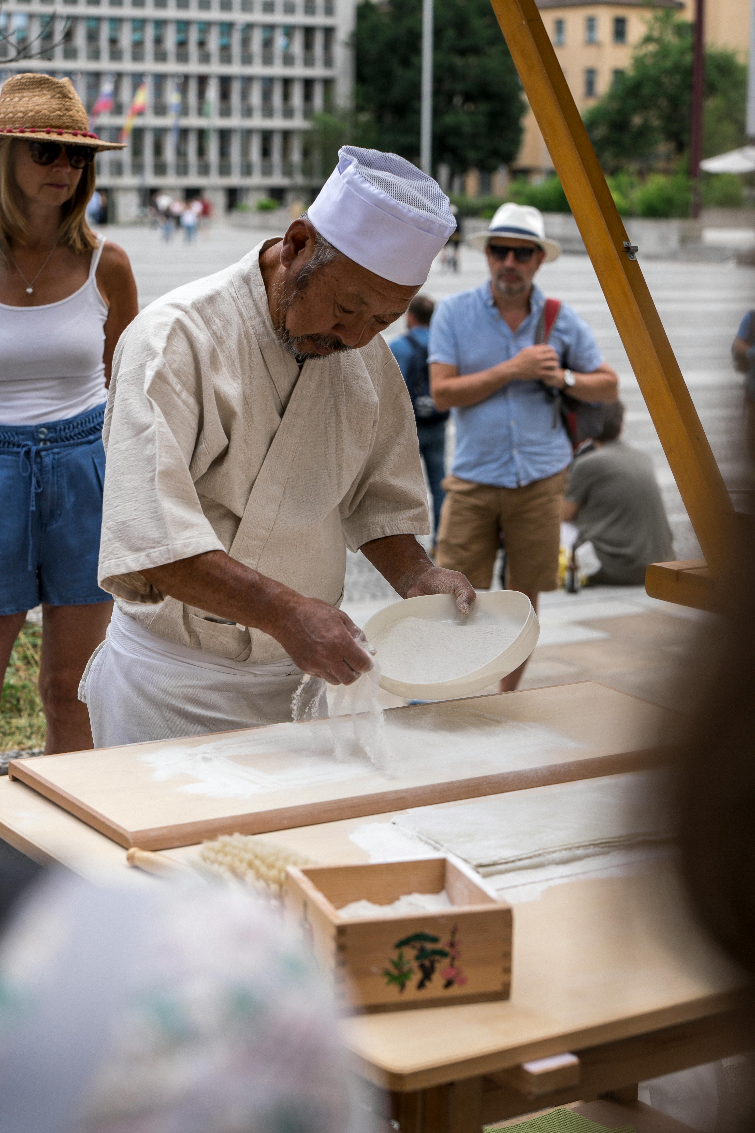 A chef in traditional attire preparing food outdoors in an urban square.